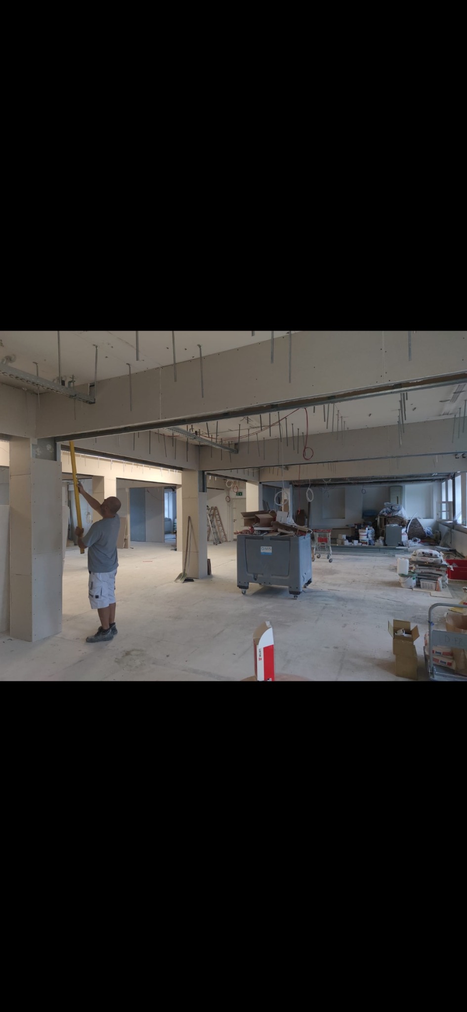 Pracownik mierzy pion żółtą poziomicą na ścianie z płyt kartonowo-gipsowych w trakcie remontu biura, widoczne metalowe profile sufitowe i narzędzia w tle.