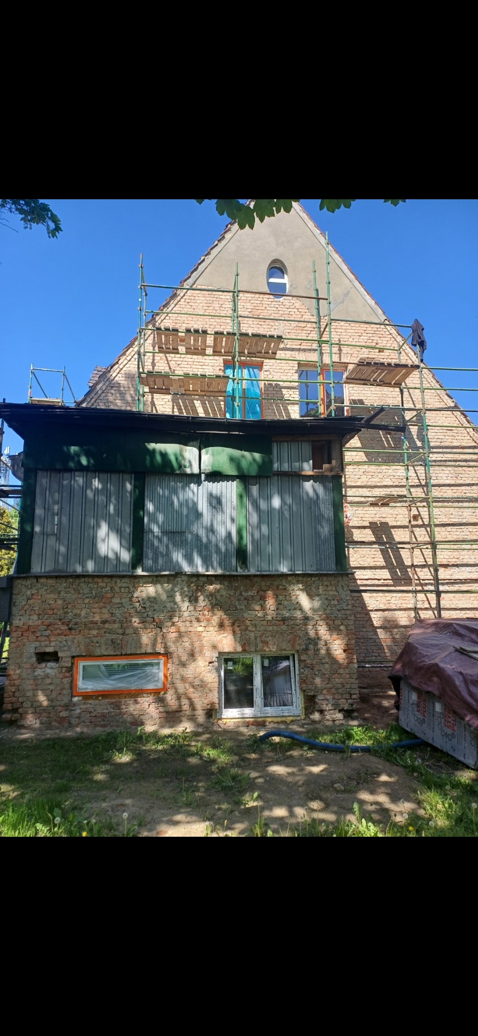 Dom z cegły w trakcie prac elewacyjnych, widoczne rusztowania, częściowo otynkowana ściana szczytowa z owalnym oknem, nowa stolarka okienna na parterze.