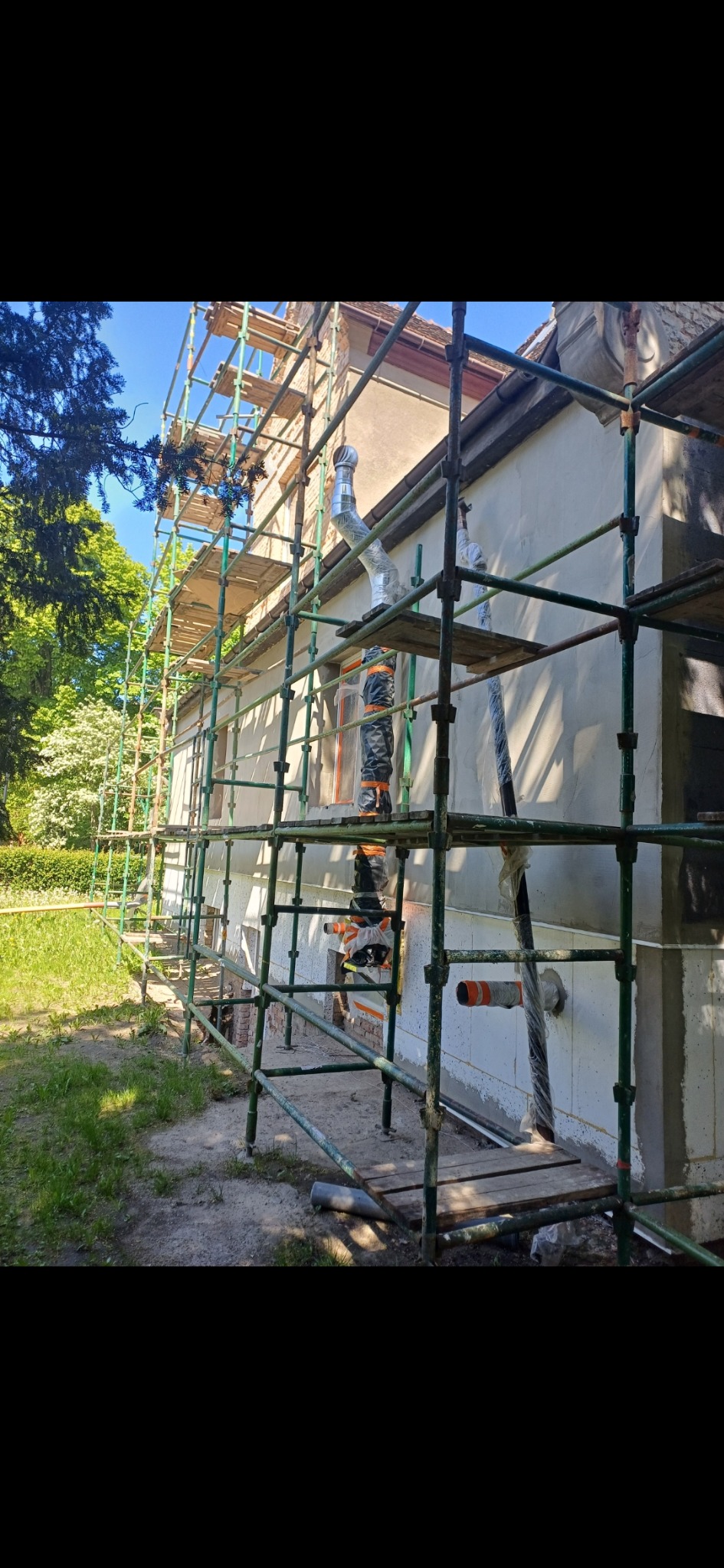 Budynek w trakcie ocieplania, widoczne rusztowania, fragment elewacji z ociepleniem, wystające rury wentylacyjne zabezpieczone folią, teren wokół budynku częściowo przygotowany pod prace.