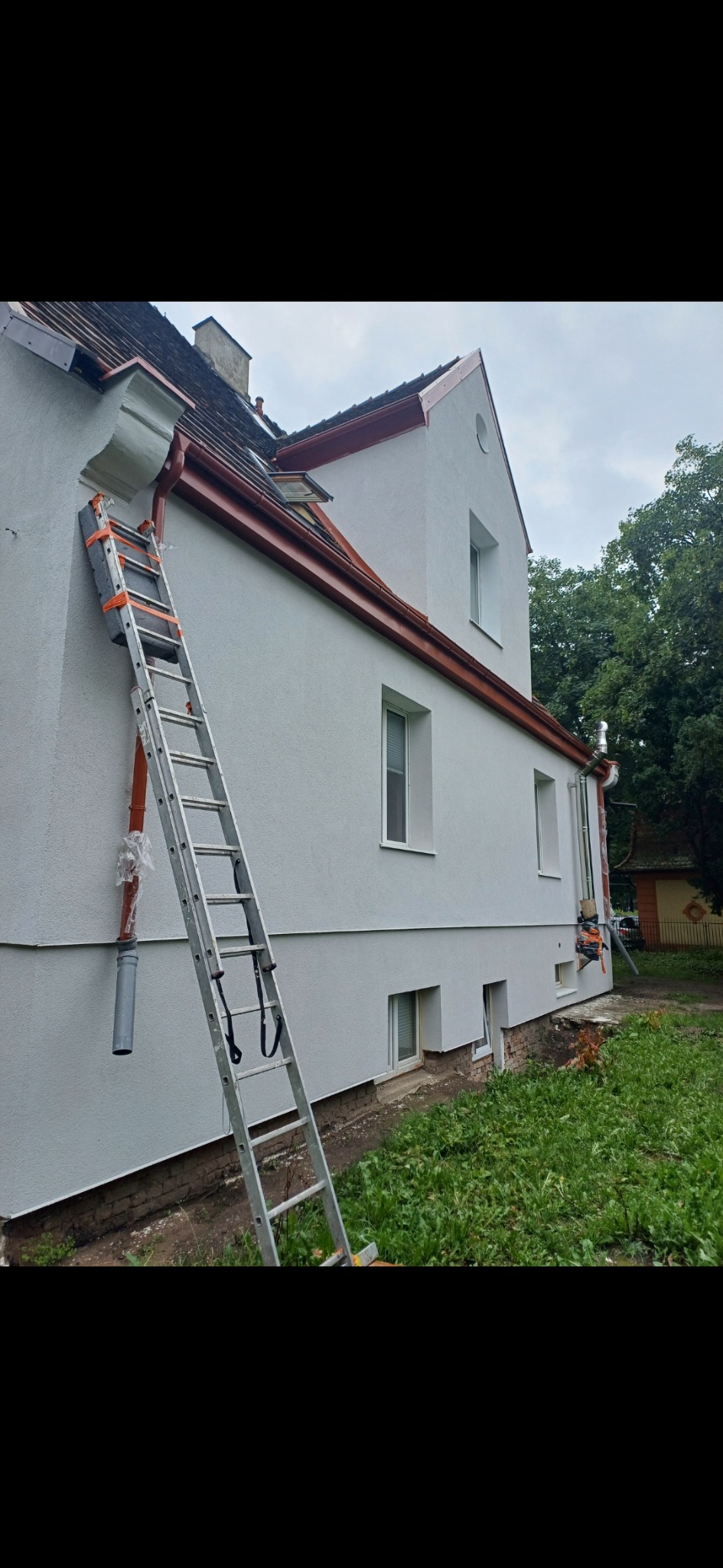 Biały dom z nową elewacją i brązowymi rynnami, z drabiną opartą o ścianę i zabezpieczeniem na rynnie.