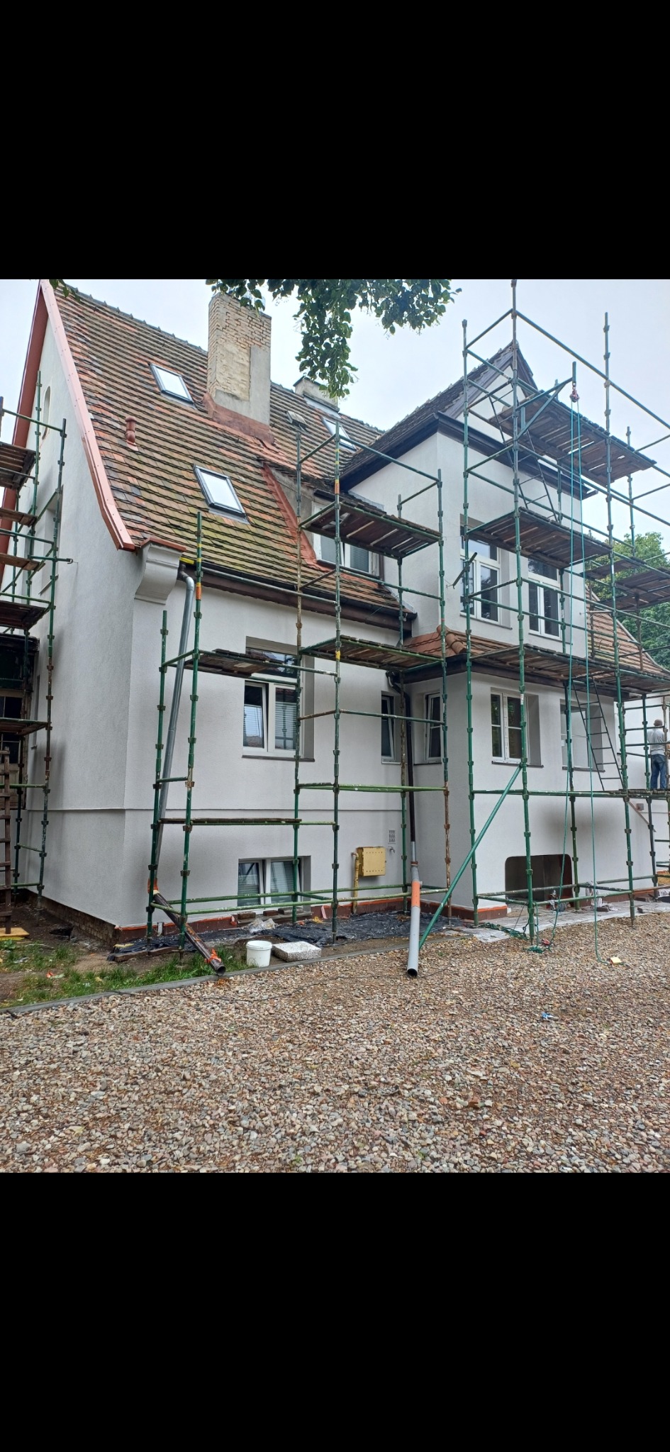 Dom jednorodzinny w trakcie wykonywania elewacji, otoczony rusztowaniem, z widoczną częściowo odnowioną dachówką i nowym tynkiem na ścianach.