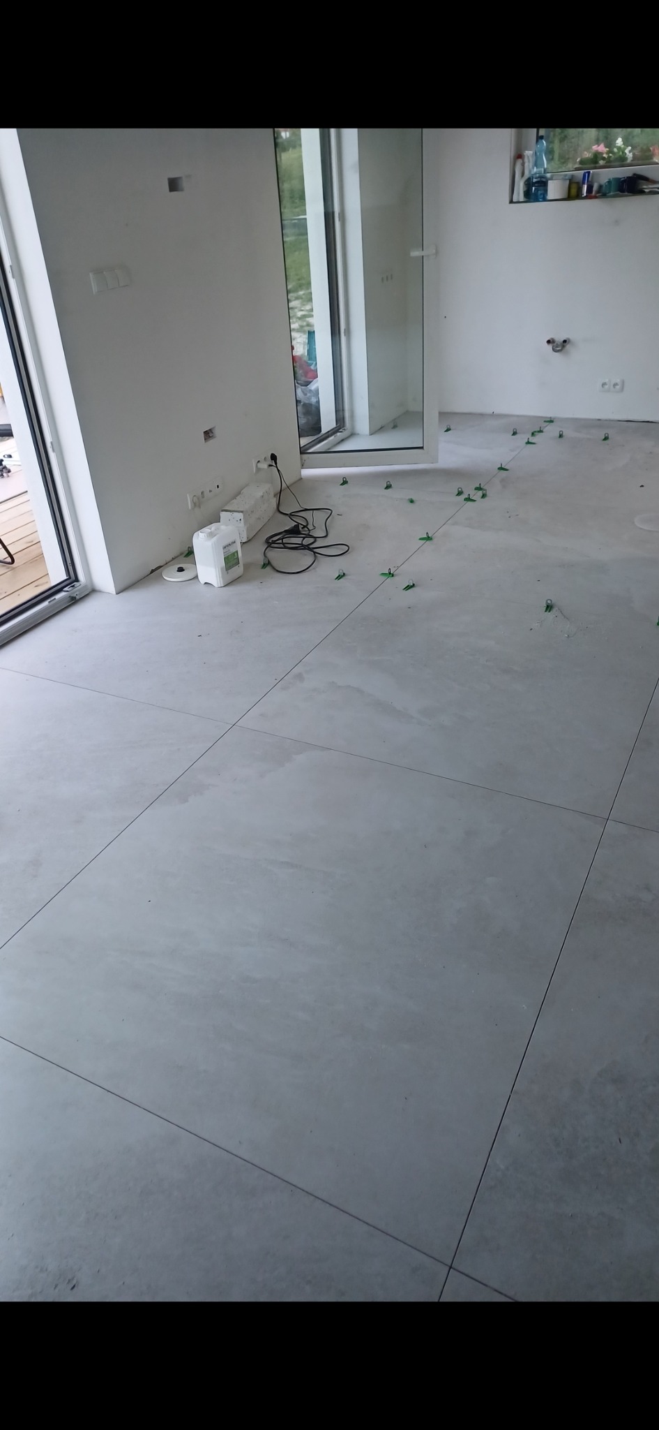 Układanie jasnych, kwadratowych płytek podłogowych z użyciem zielonych krzyżyków dystansowych w Wiślince, widoczne przygotowane materiały i przewody elektryczne.