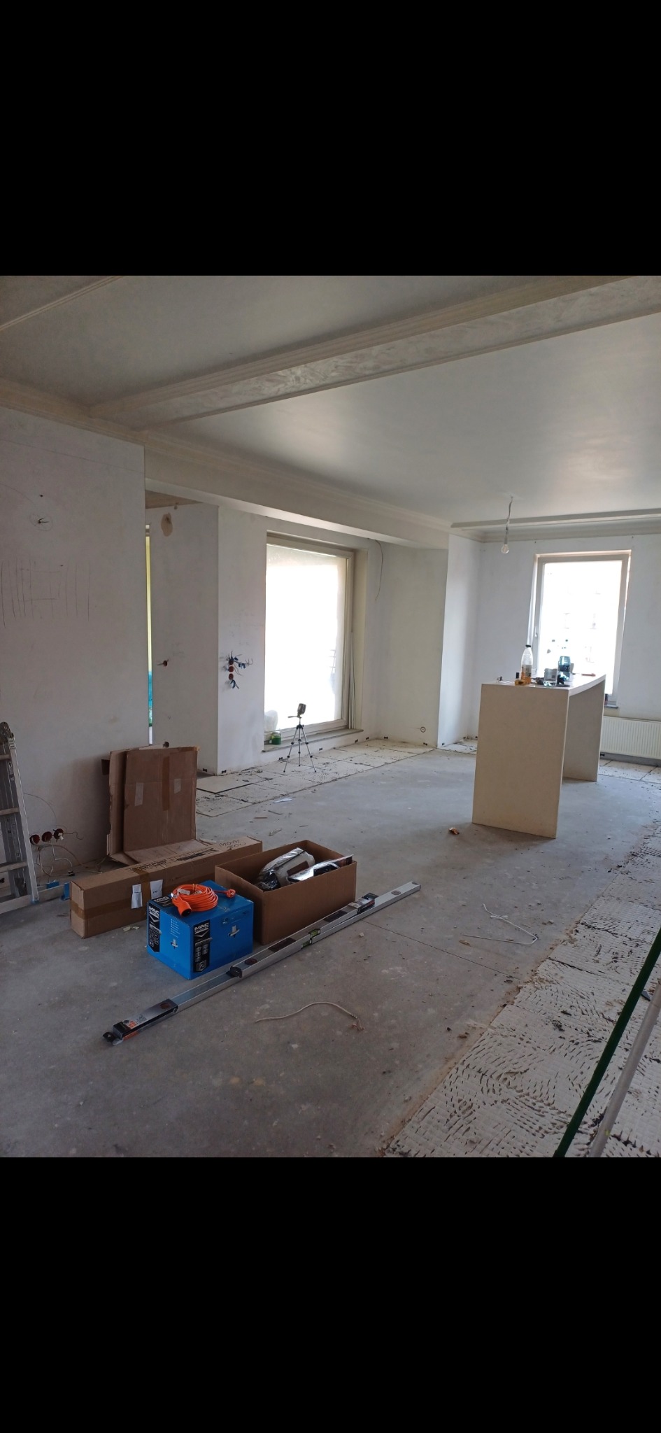 Wnętrze w trakcie prac przygotowawczych do remontu: surowe ściany, częściowo przygotowane podłoże, narzędzia i materiały budowlane w kartonach, poziomica na podłodze, widoczne ślady po instalacjach...