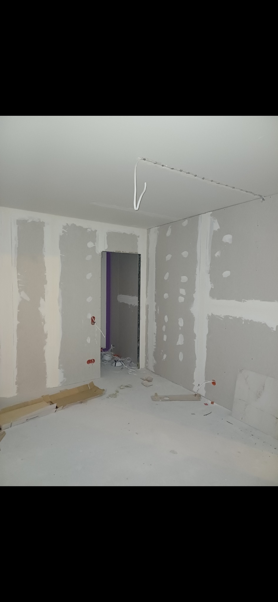 Wnętrze w trakcie prac wykończeniowych: ściany pokryte płytami kartonowo-gipsowymi z widocznymi otworami po wkrętach, surowa podłoga, wystające przewody elektryczne i fragmenty kartonów na podłodze...