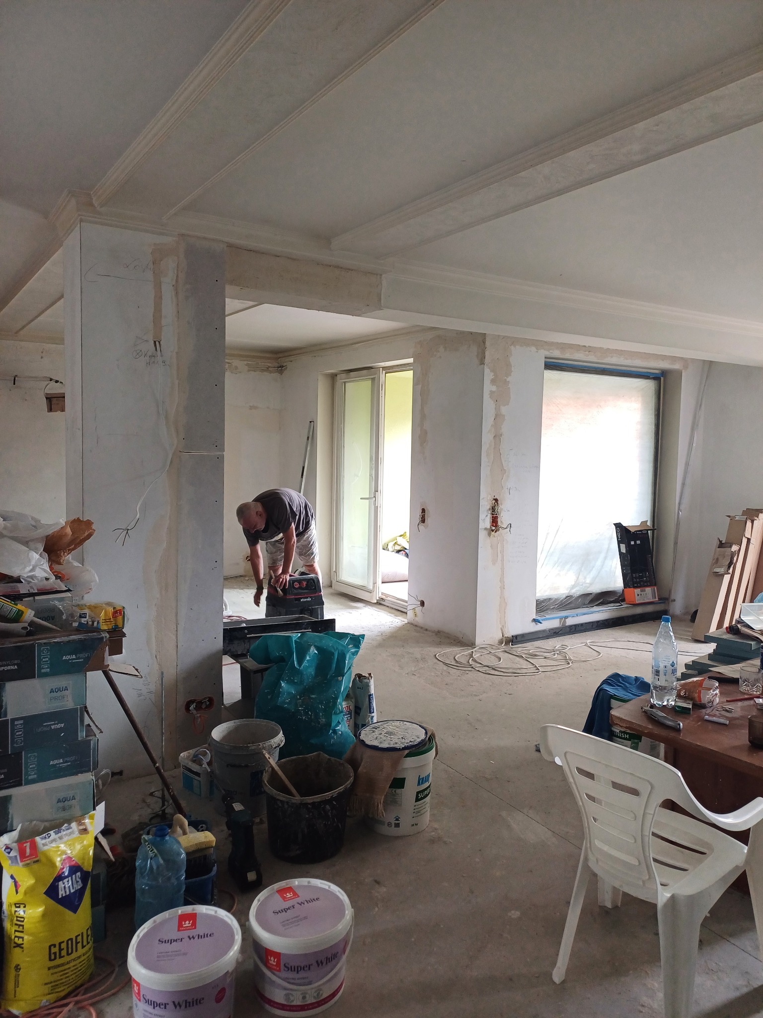 Wnętrze w trakcie prac wykończeniowych, widoczny mężczyzna szlifujący ścianę, liczne narzędzia i materiały budowlane, surowe ściany z widocznymi śladami po elektryce, listwy dekoracyjne pod sufitem...