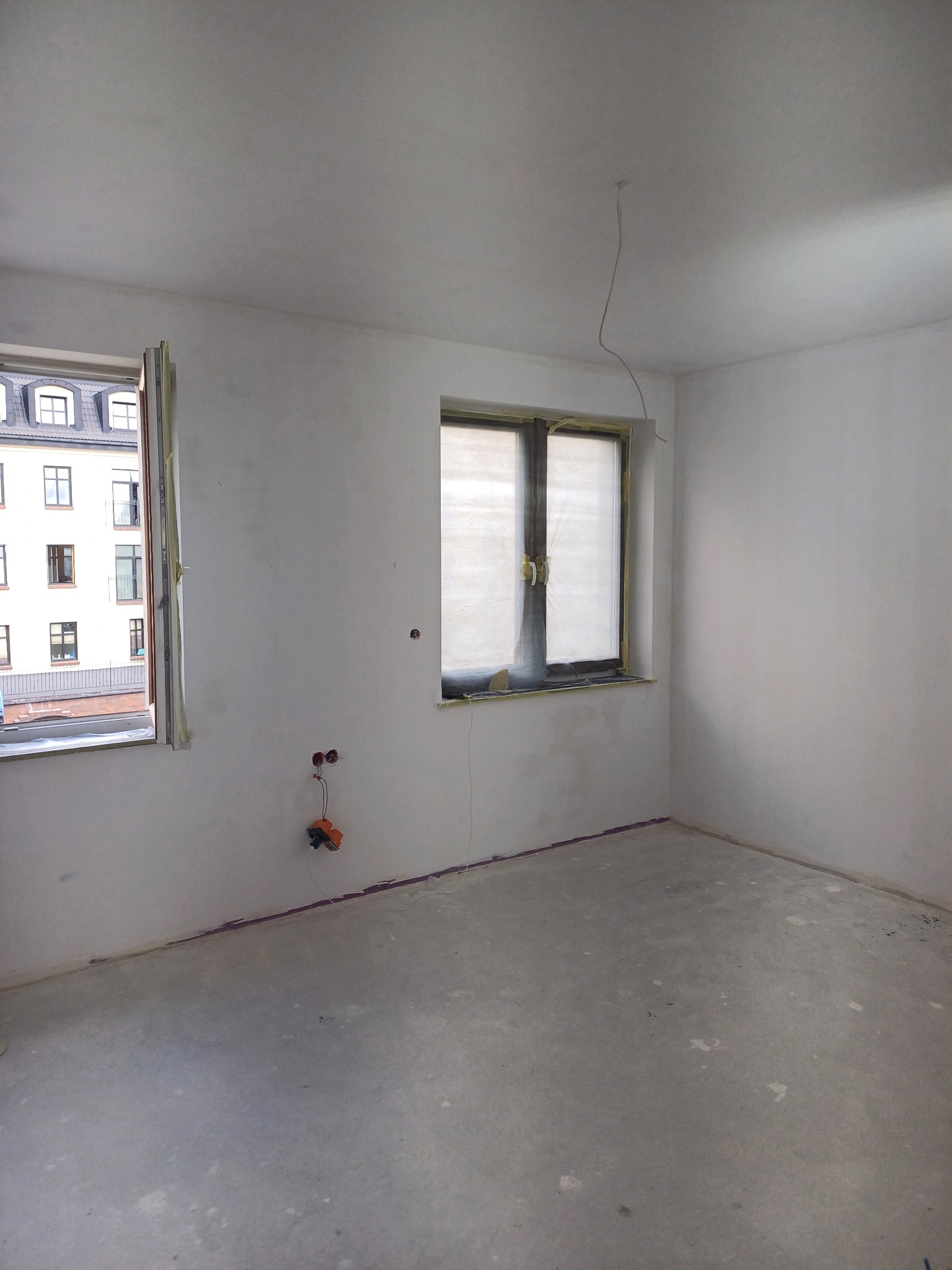 Pokój w trakcie prac wykończeniowych: ściany po nałożeniu gładzi, zabezpieczone okna folią malarską, widoczne puszki elektryczne i wystające przewody, surowa podłoga.