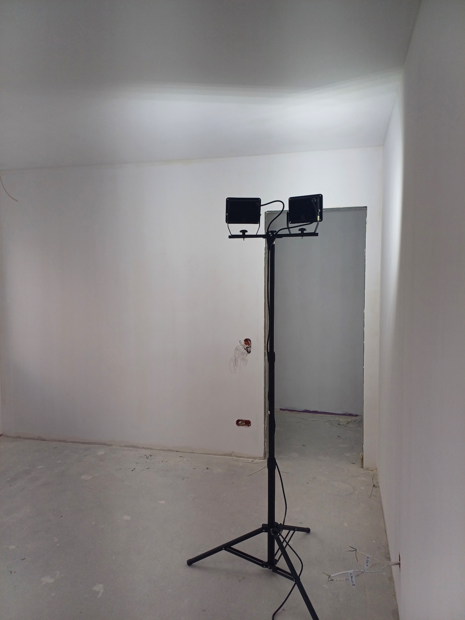 Wnętrze w trakcie malowania ścian na biało, widoczny statyw z dwoma lampami halogenowymi oświetlającymi pomieszczenie, surowy beton na podłodze, ściany po gładziowaniu, przygotowane do dalszych...