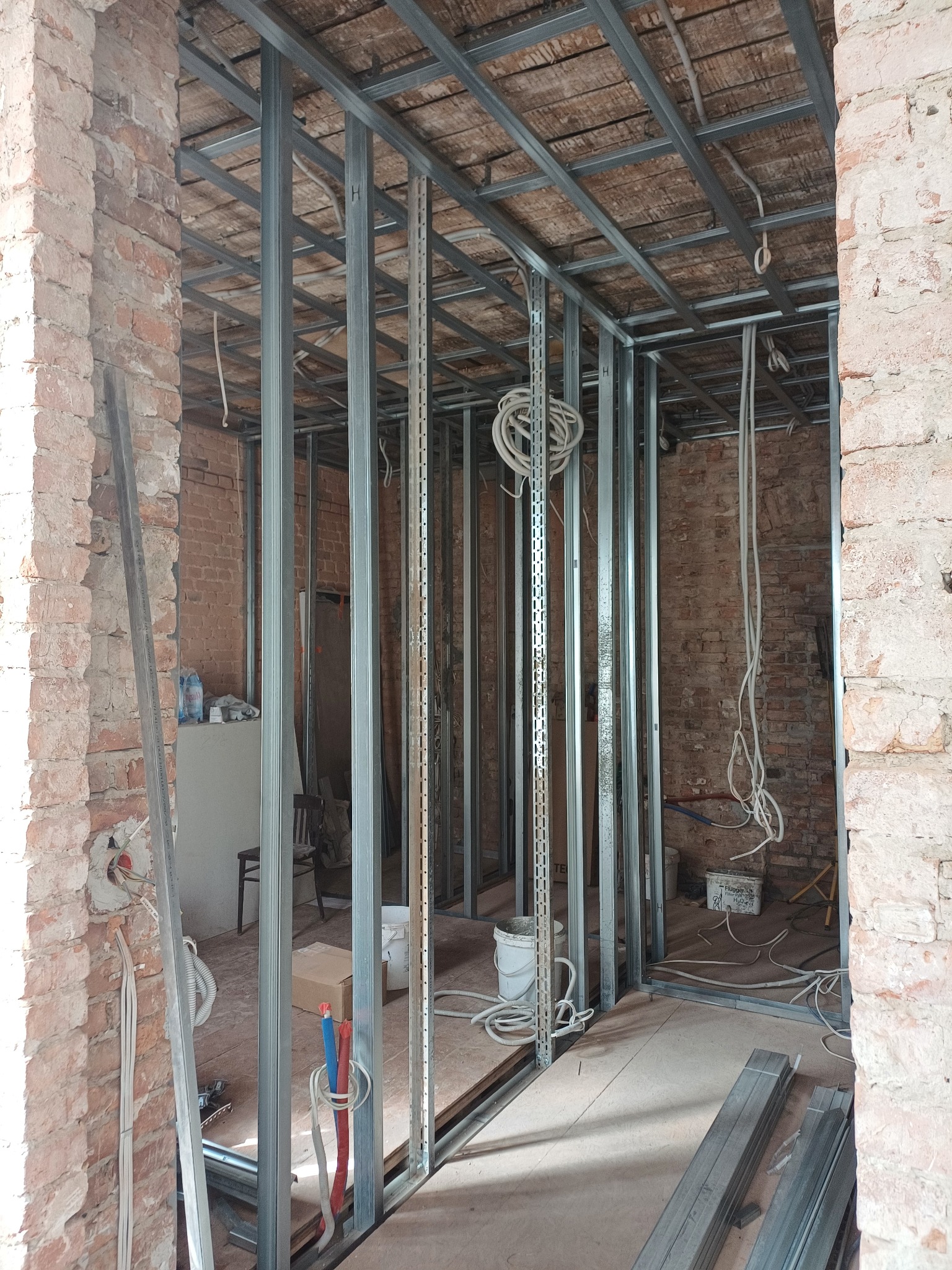 Stelaż z profili metalowych zamontowany w pomieszczeniu z surową, czerwoną cegłą, widoczne instalacje elektryczne i hydrauliczne, przygotowanie do montażu płyt gipsowo-kartonowych.
