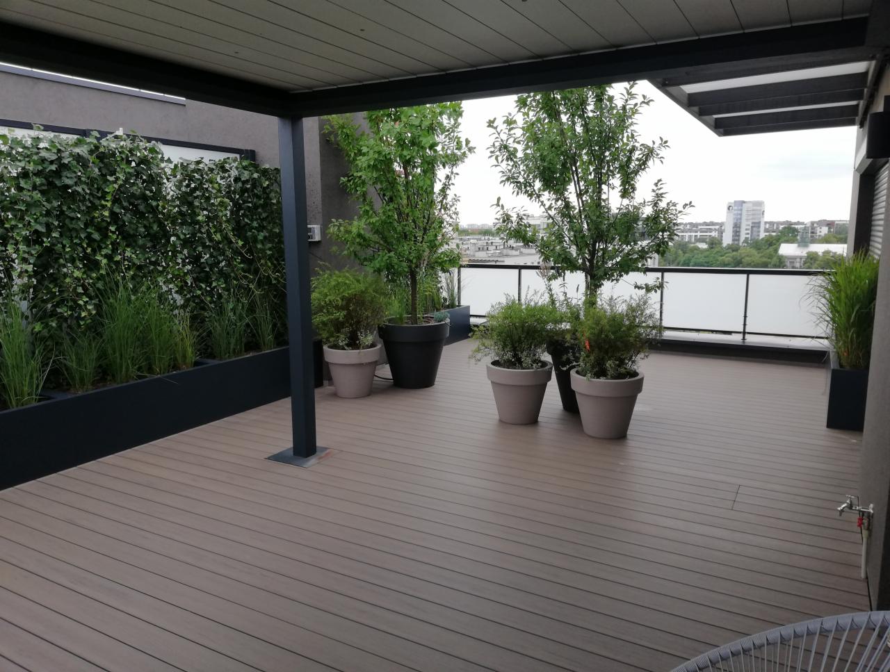 Nowoczesny taras na dachu z drewnianą podłogą, roślinami w donicach, zieloną ścianą z bluszczu i widokiem na miasto w tle.