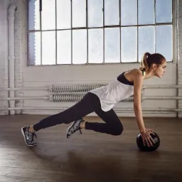 Kobieta w sportowym stroju wykonuje ćwiczenie plank z piłką lekarską w przestronnym, jasnym pomieszczeniu z drewnianą podłogą i dużymi oknami.