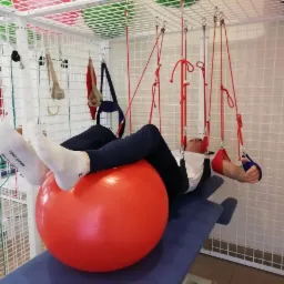 Pacjent leżący na czerwonej piłce rehabilitacyjnej, stopy w białych skarpetach Tommy Hilfiger, podwieszony na czerwonych linach w sali rehabilitacyjnej z białą kratą na ścianach i suficie.