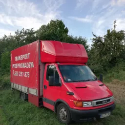 Czerwony samochód dostawczy Iveco z plandeką, na której widnieje biały napis 'TRANSPORT PRZEPROWADZKI' i numer telefonu, zaparkowany na trawie w słoneczny dzień.