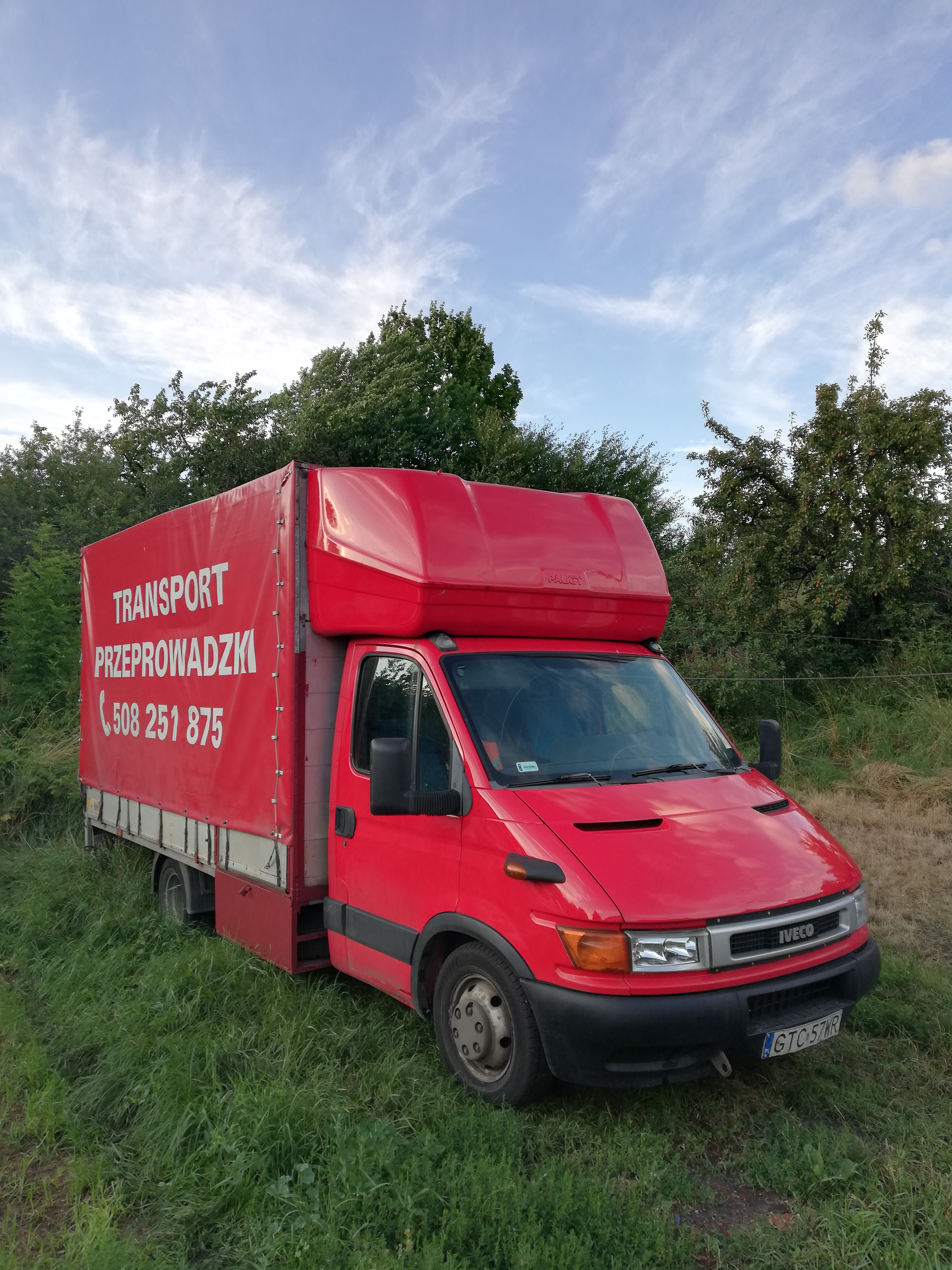 Czerwony samochód dostawczy Iveco z plandeką, na której widnieje biały napis 'TRANSPORT PRZEPROWADZKI' i numer telefonu, zaparkowany na trawie w słoneczny dzień.