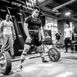 Czarno-białe zdjęcie mężczyzny przygotowującego się do podniesienia sztangi w siłowni, z dwoma obserwatorami i logotypami firm na tle.