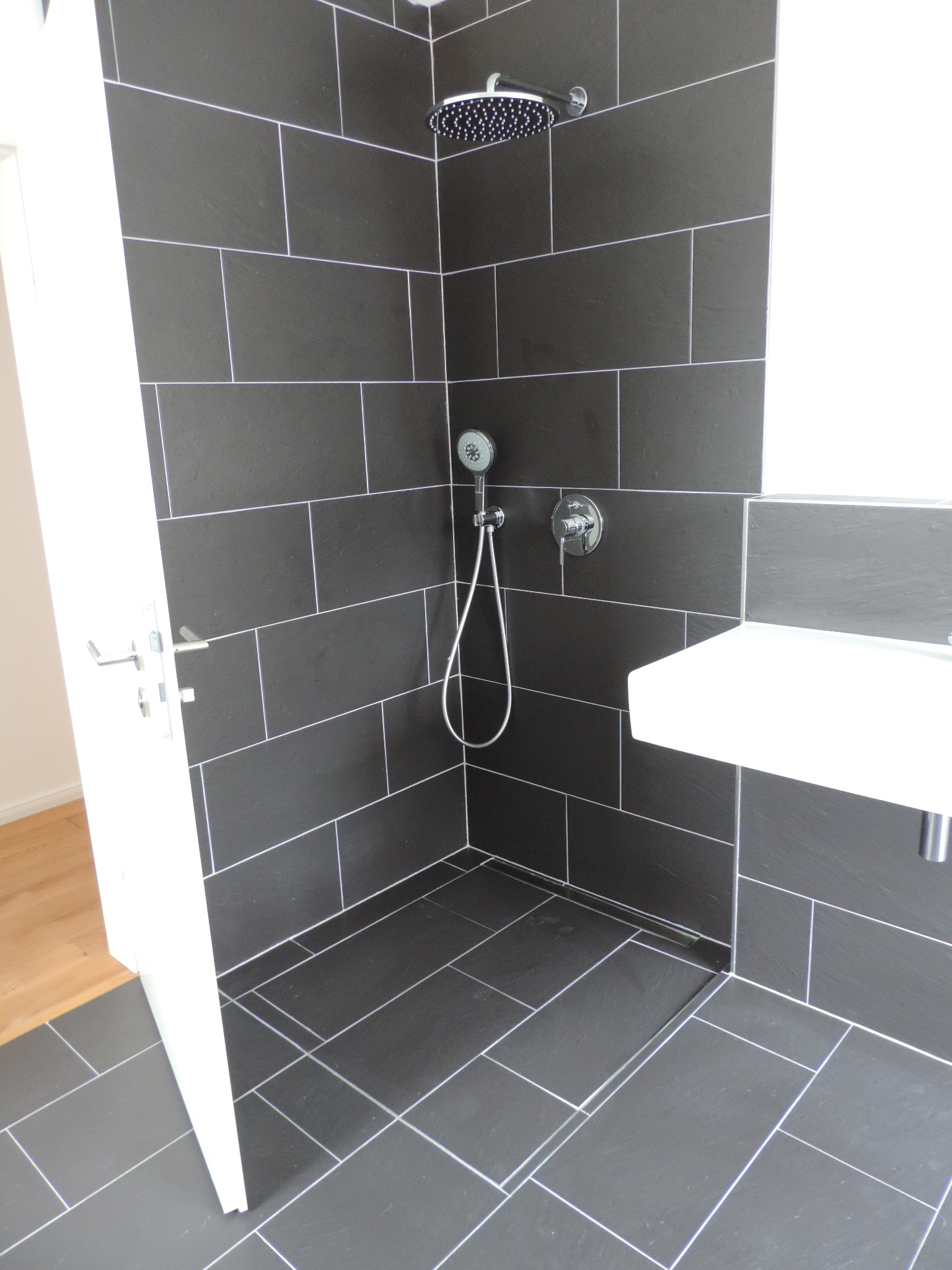 Nowoczesna kabina prysznicowa z ciemnymi, prostokątnymi płytkami i białymi fugami. Widoczny deszczownica, słuchawka prysznicowa i umywalka. Otwarta biała rama drzwi.