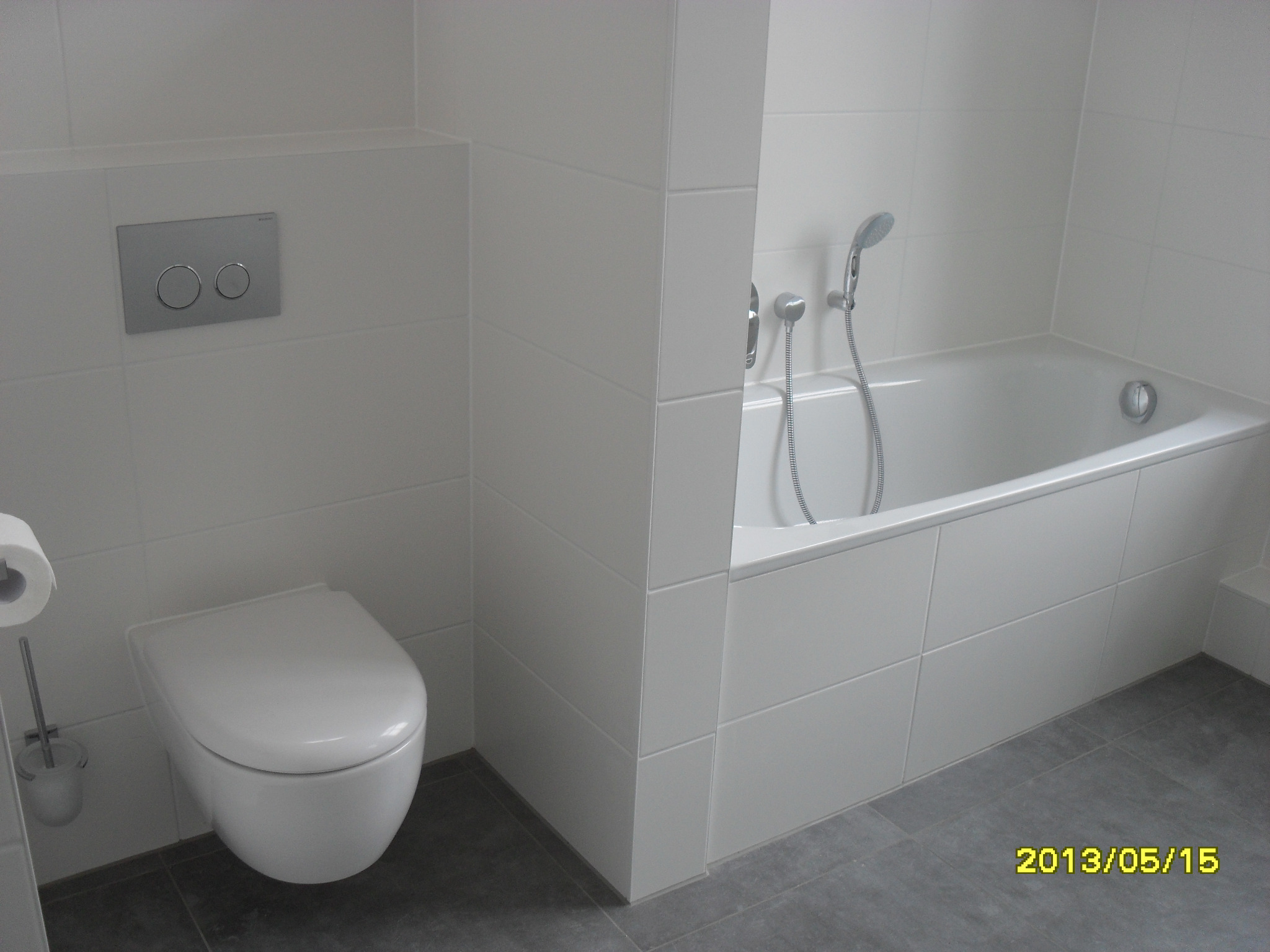 Minimalistyczna łazienka w bieli: podwieszana toaleta, wanna obudowana płytkami, szara podłoga. Widoczny fragment ściany z przyciskiem spłukującym.