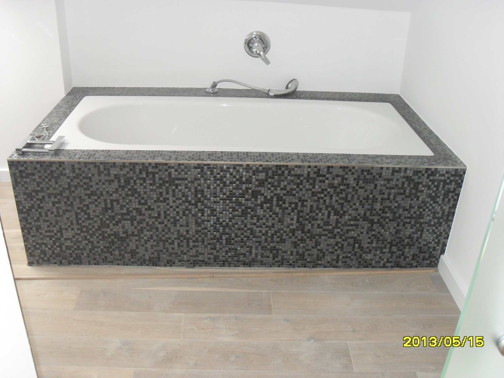 Obudowana mozaiką wanna w łazience z drewnopodobną podłogą. Bateria wannowa chromowana. Widoczny fragment kabiny prysznicowej. Ujęcie z niskiego kąta.