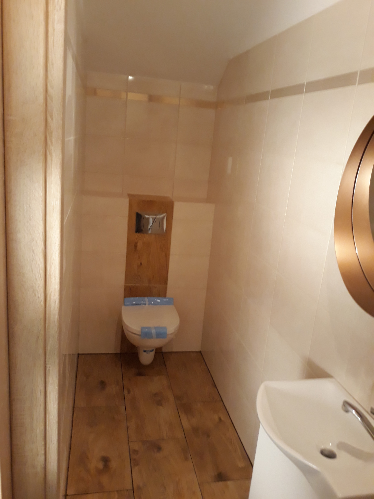 Wykończona łazienka z beżowymi płytkami, drewnopodobną podłogą i białą umywalką. Toaleta z zabezpieczeniem na desce. Okrągłe lustro z metalową ramą.