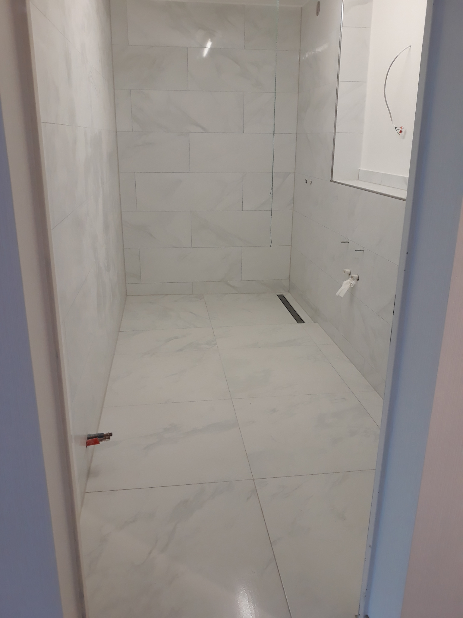 Wykończona łazienka z białymi płytkami imitującymi marmur na ścianach i podłodze. Widoczny odpływ liniowy, fragment kabiny prysznicowej i wystające rury.