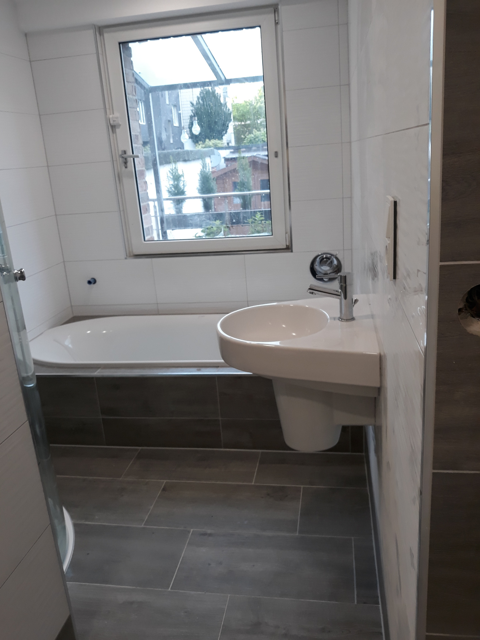 Nowoczesna łazienka z wanną, umywalką na postumencie i szarymi płytkami podłogowymi. Okno z widokiem na zewnątrz. Minimalistyczny design, jasne kolory.