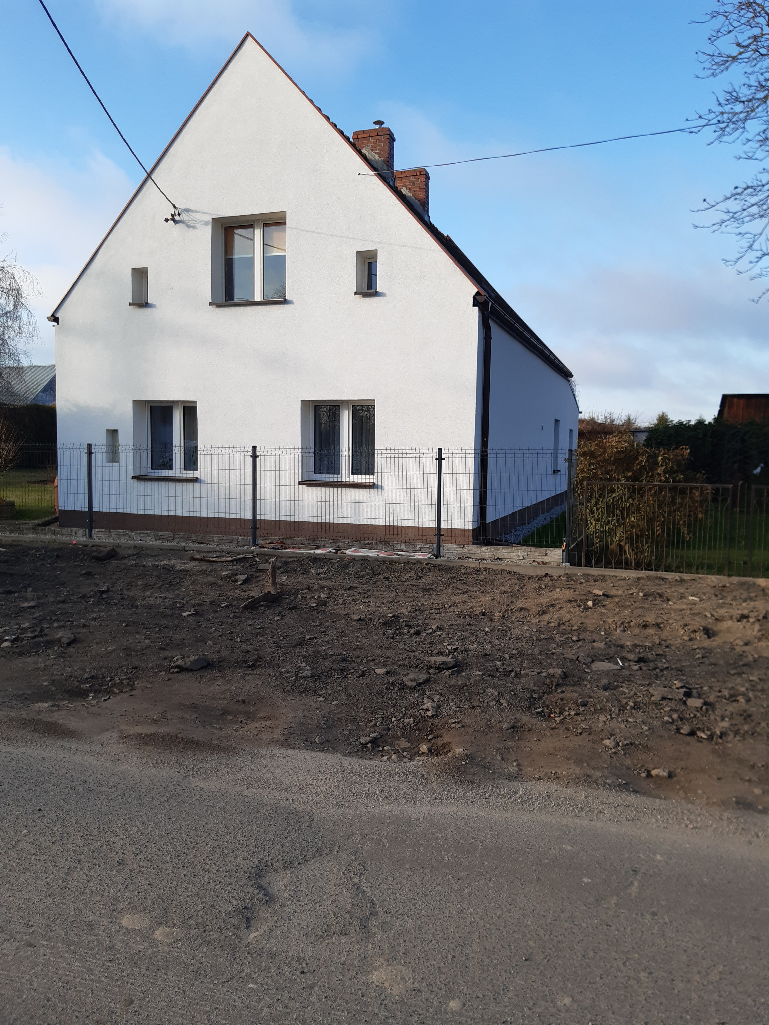 Biały dom jednorodzinny z nowym ogrodzeniem panelowym na nierównym terenie. Widok z ulicy w słoneczny dzień. Częściowo widoczne drzewa i krzewy w tle.