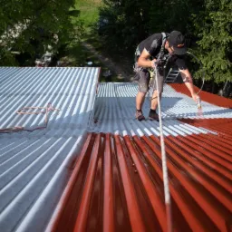 Pracownik zabezpieczony linami maluje metalowy dach na czerwono-biało za pomocą pistoletu natryskowego, widok z góry.