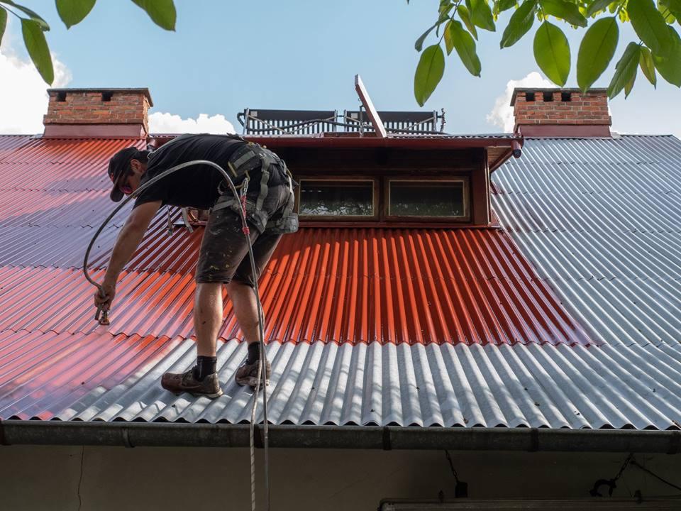 Pracownik w uprzęży maluje czerwoną farbą blaszany dach z widocznym kominem i oknem dachowym, część dachu pomalowana, część srebrna.