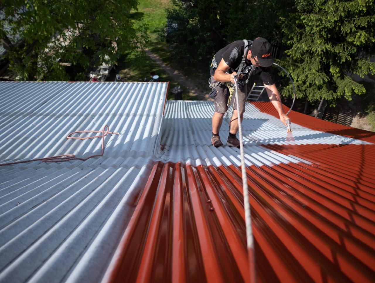 Pracownik zabezpieczony linami maluje metalowy dach na czerwono-biało za pomocą pistoletu natryskowego, widok z góry.