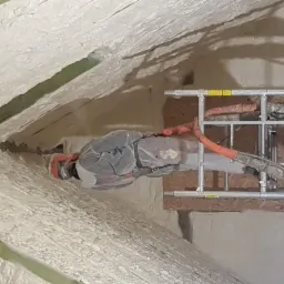 Pracownik w kombinezonie ochronnym aplikuje izolację natryskową na poddaszu, widoczny fragment rusztowania i przewody urządzenia natryskowego.