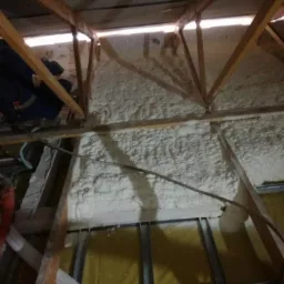Aplikacja izolacji piankowej na poddaszu widoczna z dołu, z fragmentem pracownika w roboczym ubraniu i elementami konstrukcji drewnianej oraz wentylacji.