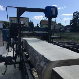 Przecieranie kłody na traku taśmowym w tartaku, widoczne budynki w tle i składowane bele siana.