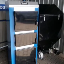 Nowy, niebiesko-biały piec z podajnikiem na ekogroszek, ustawiony na kostce brukowej w pomieszczeniu o ścianach z blachy falistej.