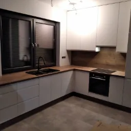 Nowoczesna kuchnia w odcieniach bieli i drewna, z czarną armaturą i oknem z żaluzjami, widziana z niskiego kąta, ukazująca minimalistyczny design i funkcjonalne rozwiązania.