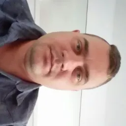 Mężczyzna w niebieskiej koszuli, z krótkimi włosami, patrzy w obiektyw. Zdjęcie portretowe, obrócone. Białe tło.