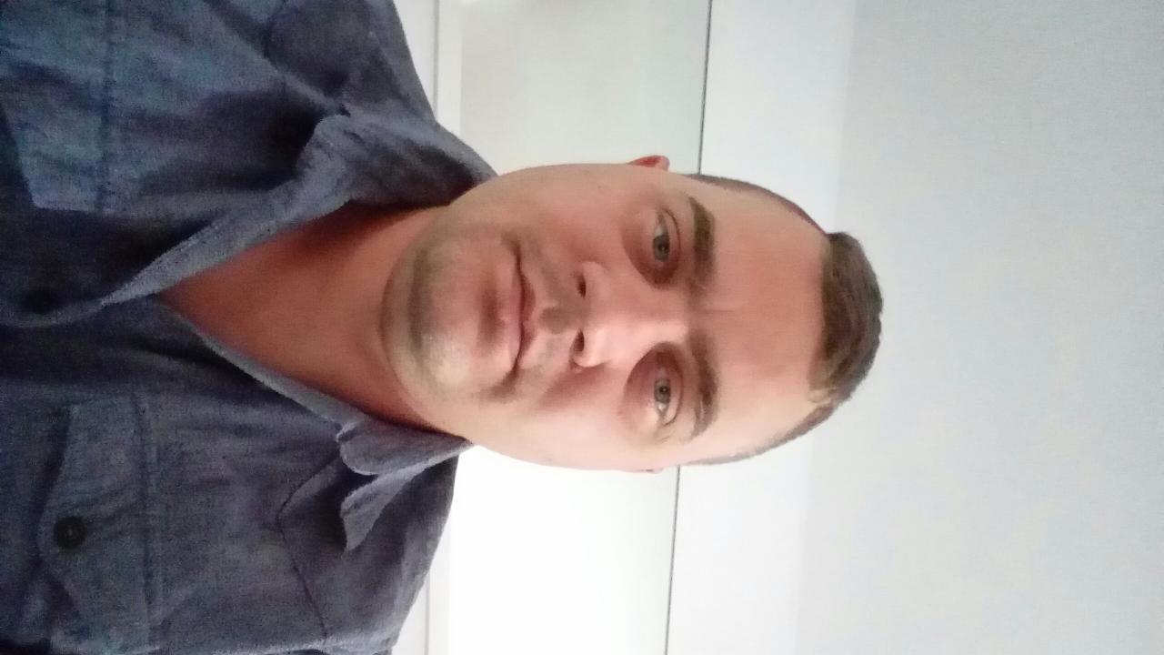 Mężczyzna w niebieskiej koszuli, z krótkimi włosami, patrzy w obiektyw. Zdjęcie portretowe, obrócone. Białe tło.