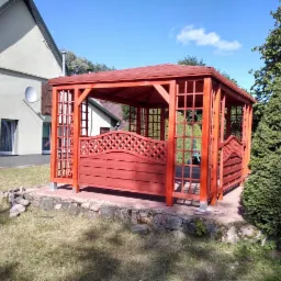 Czerwona, drewniana altana ogrodowa z dekoracyjnymi panelami kratowymi, umieszczona na podwyższeniu z kamieni, otoczona trawnikiem i zielenią.