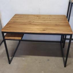 Daniel Rogala - Stół biurowy z blatem z jasnego drewna i czarną, metalową ramą, widoczny z perspektywy lekko z góry, z niewielką półką pod blatem po lewej stronie.