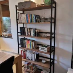 Metalowy regał magazynowy z drewnianymi półkami, wypełniony książkami, dekoracjami i pudełkami, ustawiony w jasnym pomieszczeniu obok drewnianego stołu.