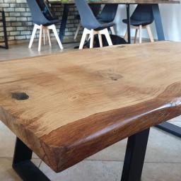 Daniel Rogala - Stół z blatem z litego drewna o nieregularnych krawędziach, wsparty na czarnych, metalowych nogach w kształcie litery V, widoczny w nowoczesnym wnętrzu z ceglaną ścianą w tle.