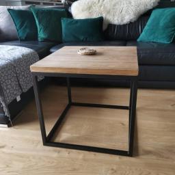 Daniel Rogala - Stolik kawowy z drewnianym blatem i czarną metalową ramą, ustawiony na jasnej drewnianej podłodze, z fragmentem czarnej skórzanej sofy i zielonymi poduszkami w tle.