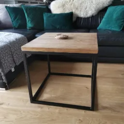 Stolik kawowy z drewnianym blatem i czarną metalową ramą, ustawiony na jasnej drewnianej podłodze, z fragmentem czarnej skórzanej sofy i zielonymi poduszkami w tle.