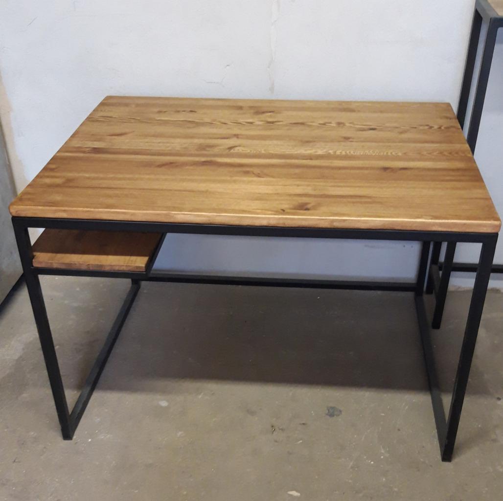 Stół biurowy z blatem z jasnego drewna i czarną, metalową ramą, widoczny z perspektywy lekko z góry, z niewielką półką pod blatem po lewej stronie.