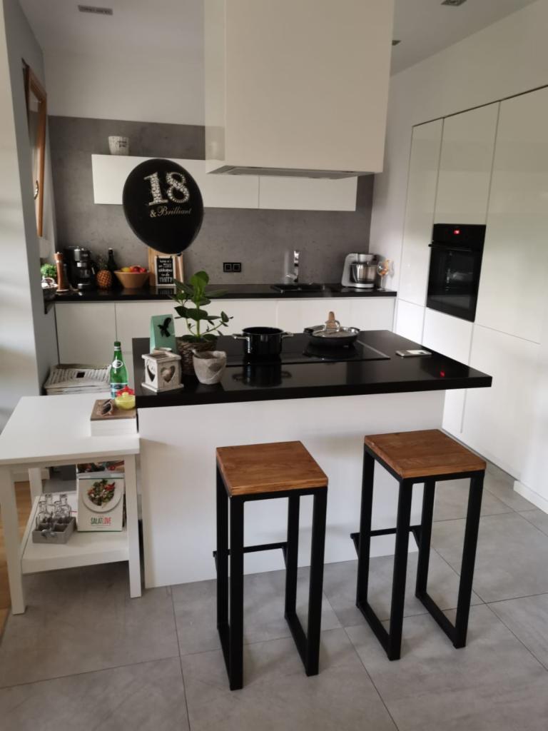 Nowoczesna kuchnia z wyspą, czarnym blatem, białymi szafkami i dwoma drewnianymi hokerami z czarnymi nogami. Na wyspie dekoracje, w tle czarny balon z napisem '18 & Brilliant'.