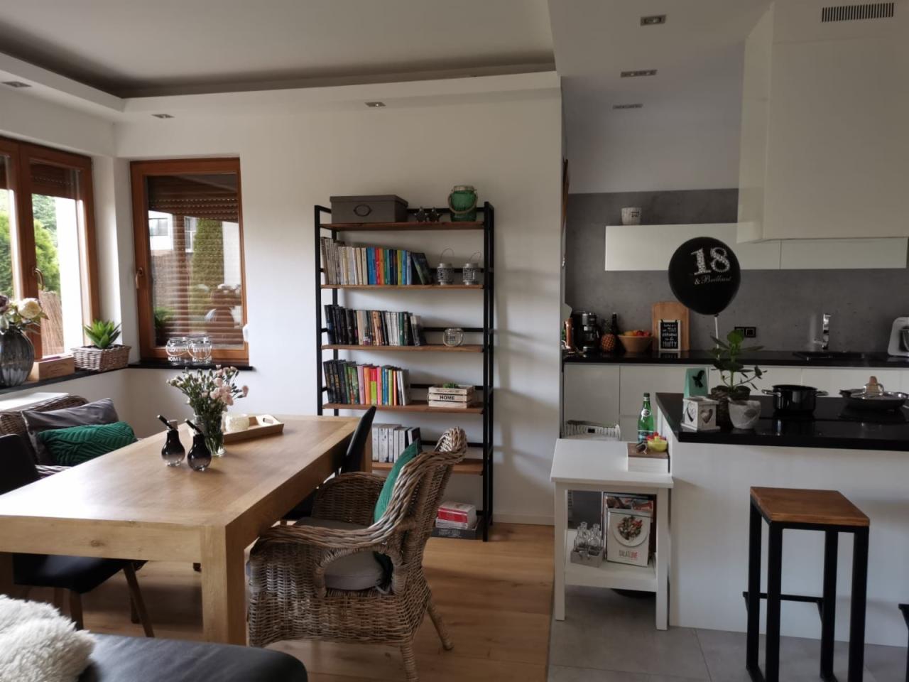 Wnętrze domu z jadalnią i kuchnią: drewniany stół, wiklinowe krzesło, regał na książki z metalową ramą, czarny balon z napisem '18' w tle.