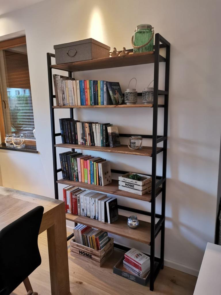 Metalowy regał magazynowy z drewnianymi półkami, wypełniony książkami, dekoracjami i pudełkami, ustawiony w jasnym pomieszczeniu obok drewnianego stołu.