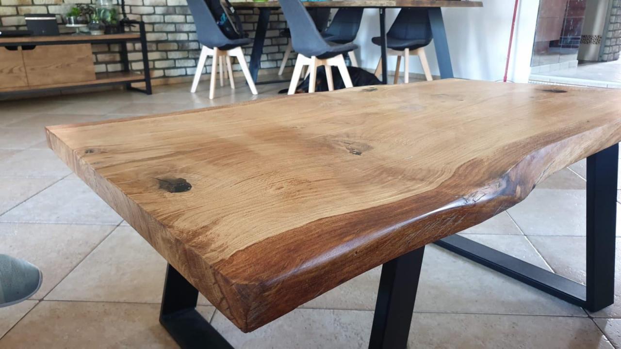 Stół z blatem z litego drewna o nieregularnych krawędziach, wsparty na czarnych, metalowych nogach w kształcie litery V, widoczny w nowoczesnym wnętrzu z ceglaną ścianą w tle.