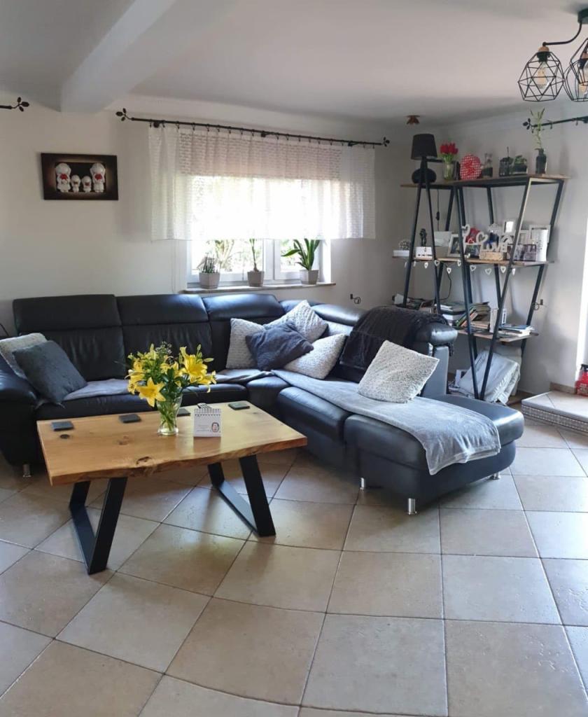 Salon z narożną, czarną sofą, drewnianym stolikiem kawowym z metalowymi nogami w kształcie litery V, regałem z metalową ramą i drewnianymi półkami oraz kwiatami w wazonie.