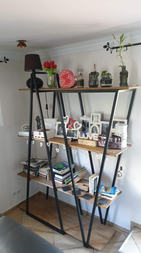 Trzypoziomowy regał o konstrukcji stalowej z drewnianymi półkami, wypełniony książkami, ramkami na zdjęcia i dekoracjami w stylu domowym, ustawiony przy białej ścianie.