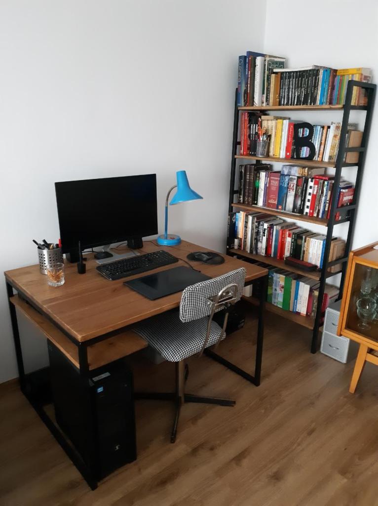 Drewniane biurko z czarną metalową ramą, komputerem, tabletem graficznym i niebieską lampką, obok regał z książkami z czarną ramą i drewnianymi półkami, krzesło w pepitkę.