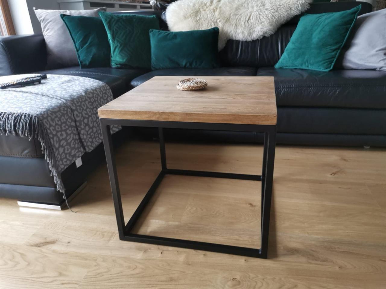 Stolik kawowy z drewnianym blatem i czarną metalową ramą, ustawiony na jasnej drewnianej podłodze, z fragmentem czarnej skórzanej sofy i zielonymi poduszkami w tle.